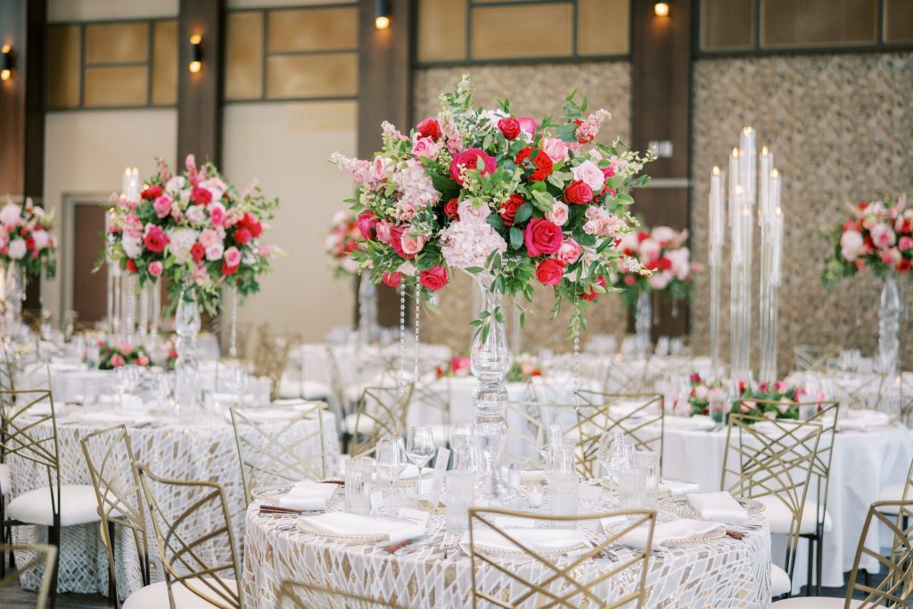 Elegant wedding reception setup in Columbus, Ohio featuring tall floral centerpieces with pink and red blooms, gold geometric chairs, white patterned linens, crystal candle holders, and an upscale ballroom reception space.