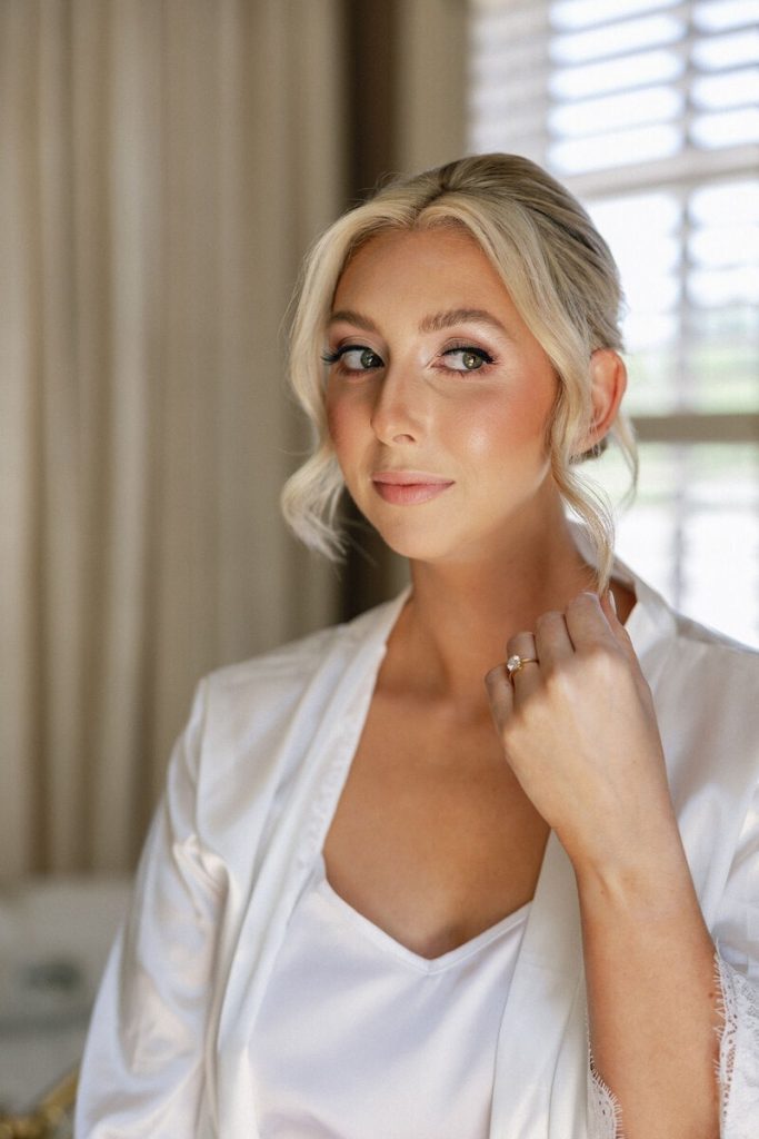 Bride with soft glam wedding makeup and a low, loosely styled updo getting ready on her wedding day in Columbus, Ohio. She wears a white bridal robe and satin camisole while posing near a window with natural light.