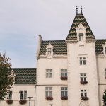 Historic European castle-style building with decorative patterned roof tiles, flower-filled window boxes, and elegant architectural details.