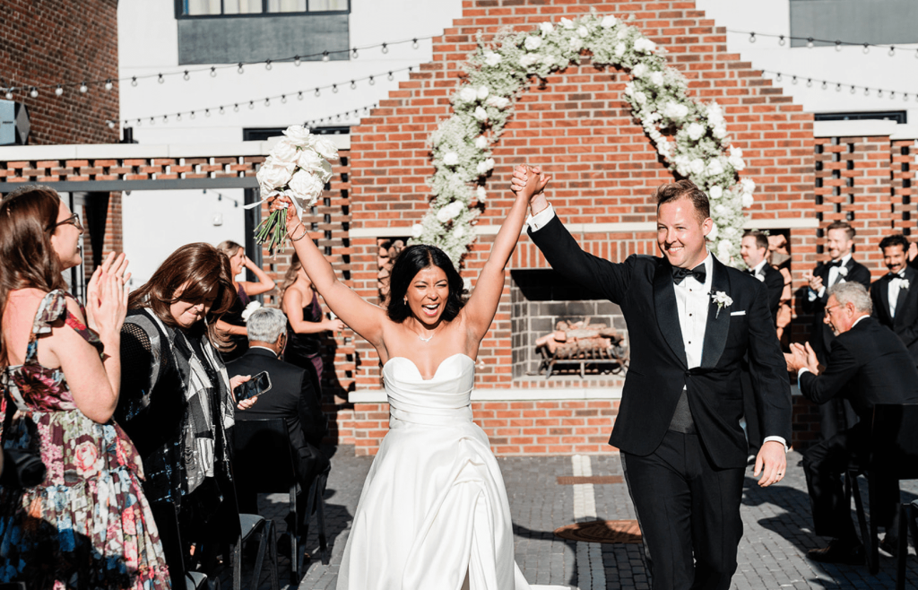 Newly married couple celebrating their ceremony recessional at The Junto in downtown Columbus, Ohio, walking beneath a floral arch surrounded by guests during an outdoor wedding ceremony.