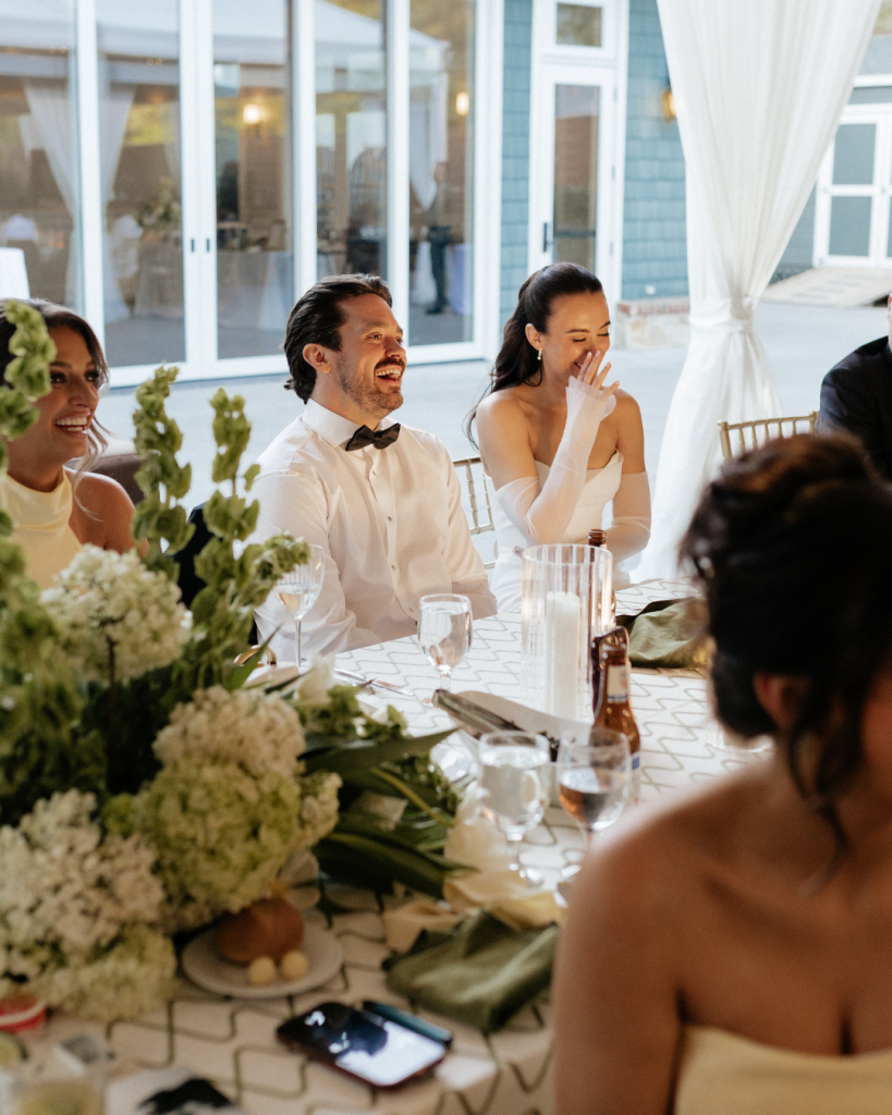 Bride and groom laugh during heartfelt reception speeches surrounded by lush spring floral arrangements.