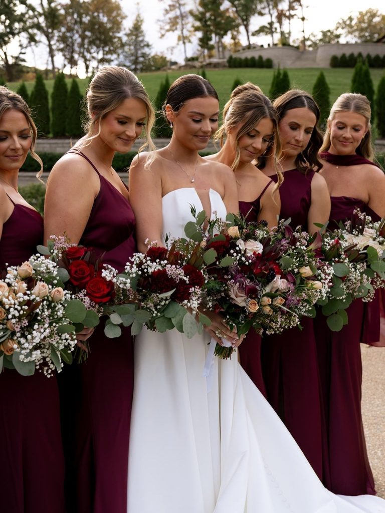 Bride with her bridesmaids in deep red dresses posing in the garden space at Knotting Hills holding fall themed bouquets from Blissful Bloom by Becky.