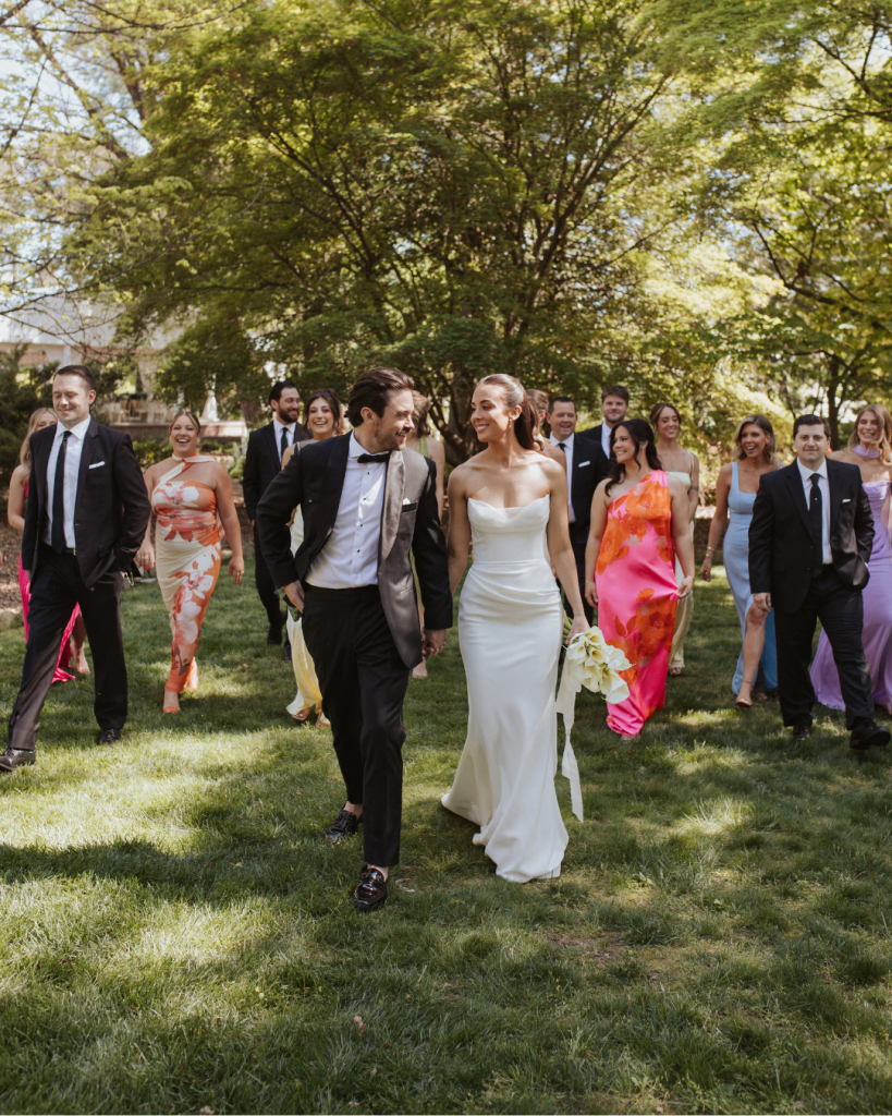 Bride and groom walk hand in hand with their wedding party through the gardens of The VanLandingham Estate following their ceremony in Charlotte.