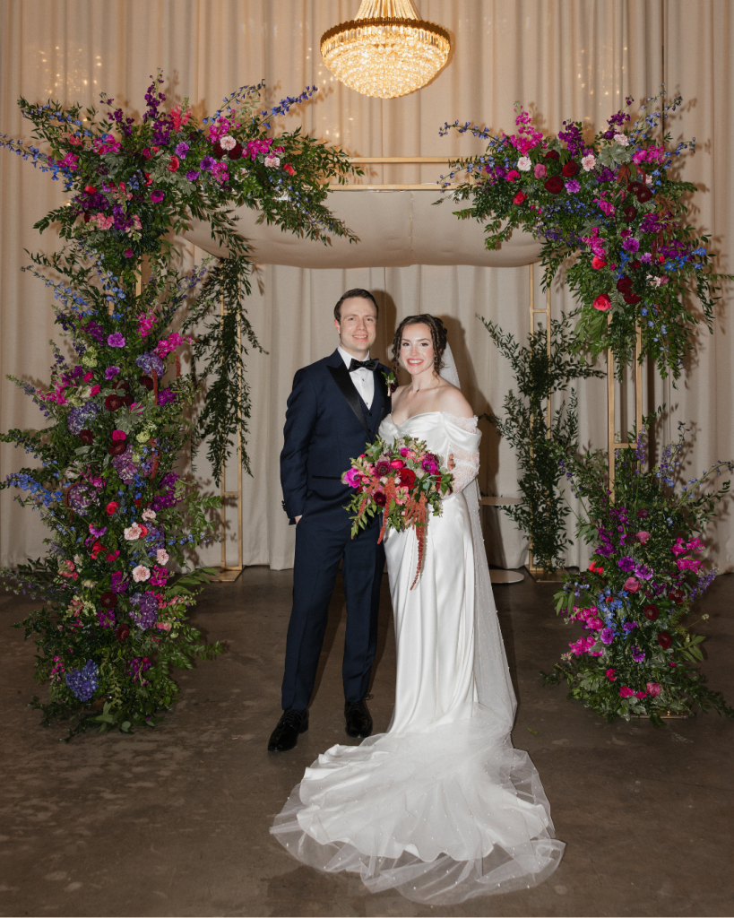 Bride and groom stand beneath a dramatic floral chuppah during their wedding reception at The Casey in Charlotte, surrounded by colorful floral installations.