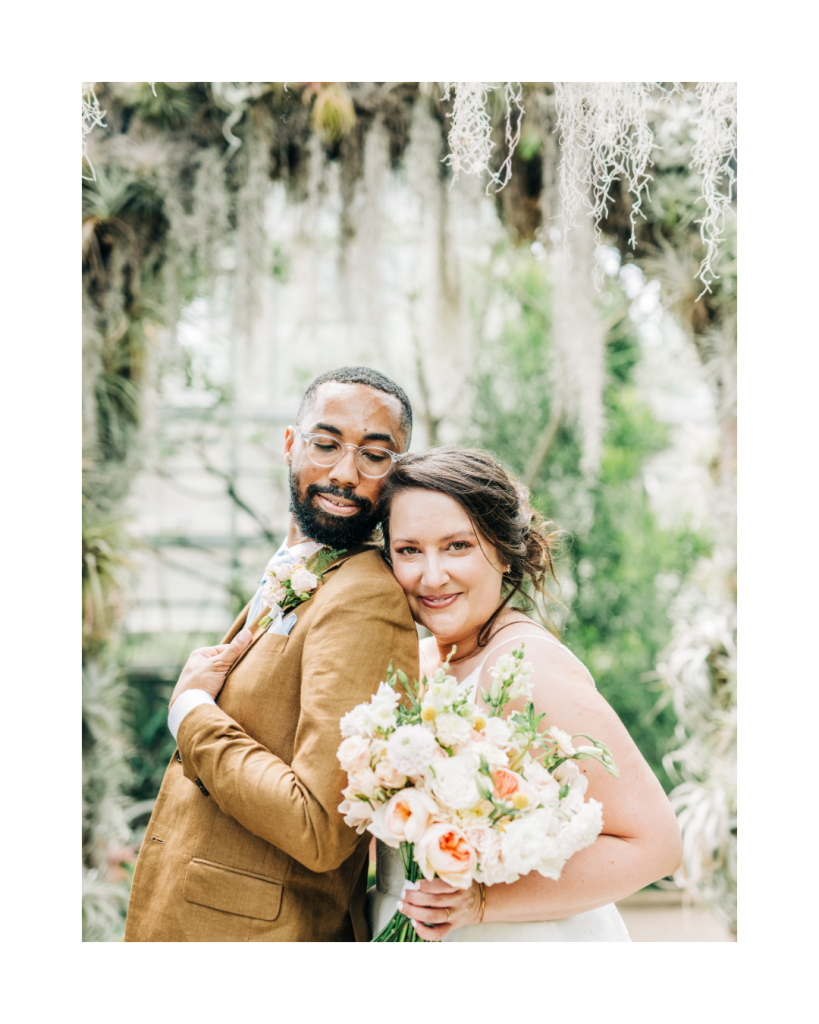 Bride and groom embrace inside the conservatory with a bouquet of blush, peach, and white flowers. Romantic wedding portrait photographed by Caitlin Elizabeth Photography for Wed Society Charlotte.