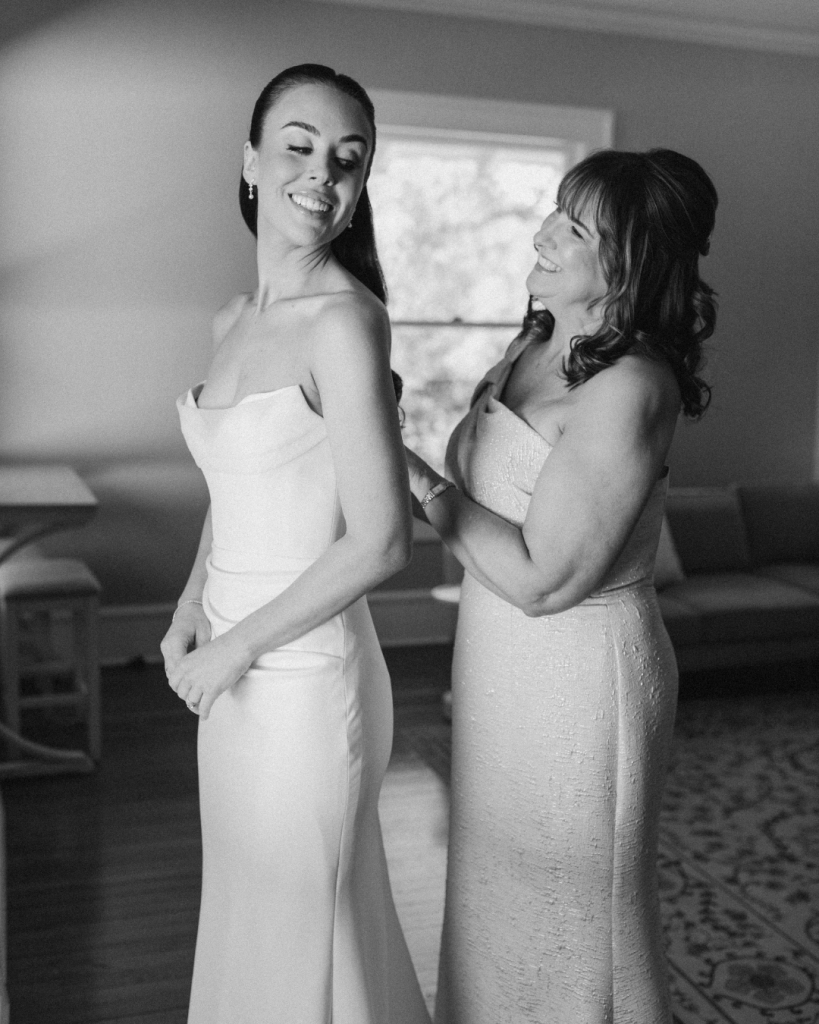 Bride smiles as her mother helps fasten her elegant wedding gown during bridal preparations before the ceremony.