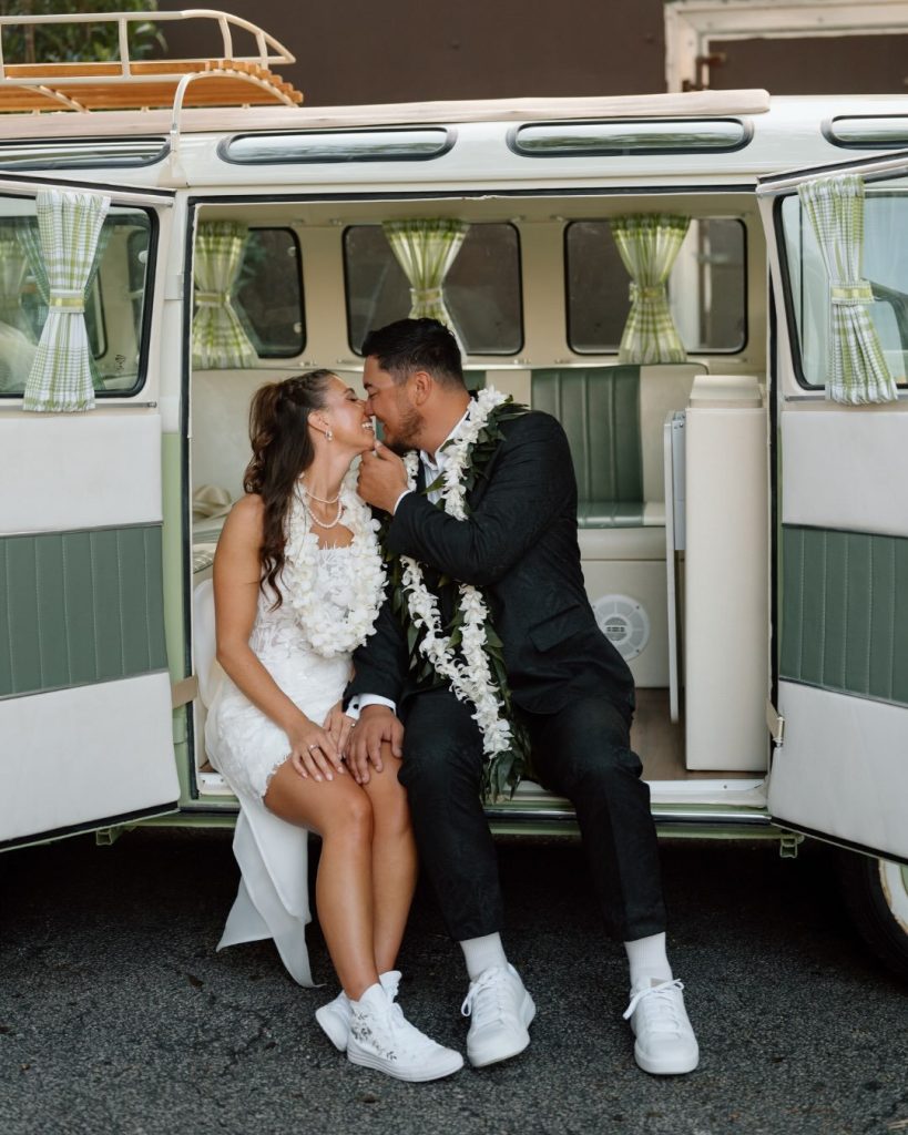 Newlyweds wearing Hawaiian lei garlands sitting together in the back of an open vintage sage green VW bus.