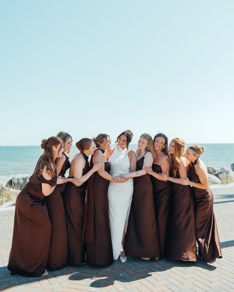 Bride standing arm in arm with bridesmaids in brown dresses, lined up along a beachfront walkway.
