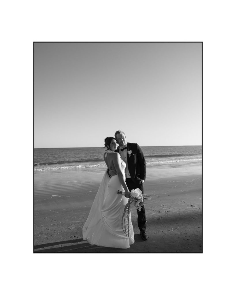 Black-and-white photo of bride and groom embracing on the beach shoreline.