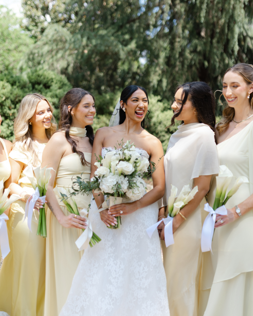 Bride laughs with bridesmaids holding white calla lily bouquets during outdoor wedding portraits at The VanLandingham Estate in Charlotte.