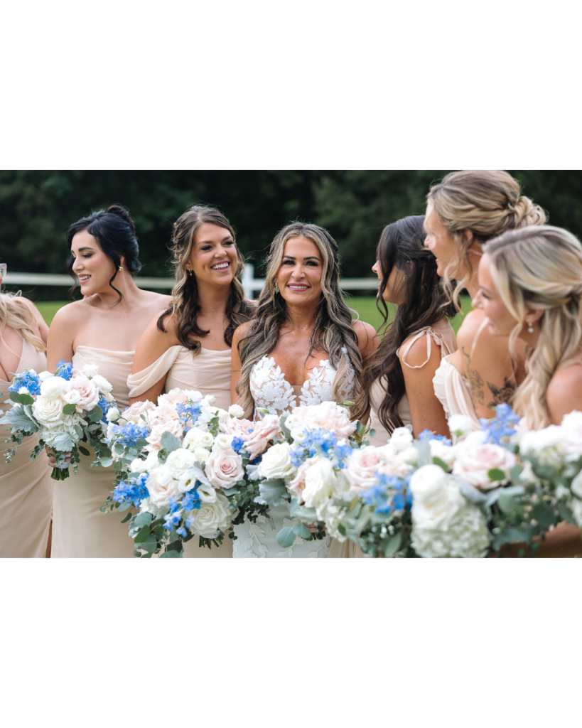 Bride stands with bridesmaids holding lush bouquets filled with blush roses, white blooms, eucalyptus, and blue delphinium during outdoor portraits.