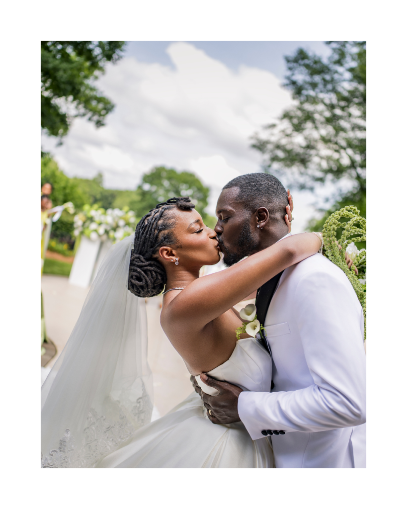 Bride and groom share their first kiss during the outdoor ceremony beneath lush floral arrangements overlooking the water.
