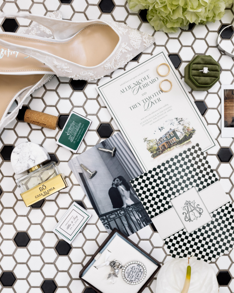 Flat lay featuring wedding invitations, rings, perfume, heirloom jewelry, and stationery with green and black details.