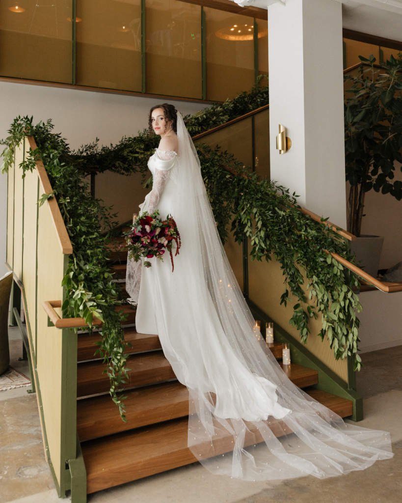 Bride poses on a staircase surrounded by cascading greenery while wearing an off the shoulder satin wedding gown and cathedral veil at the Grand Bohemian Hotel.