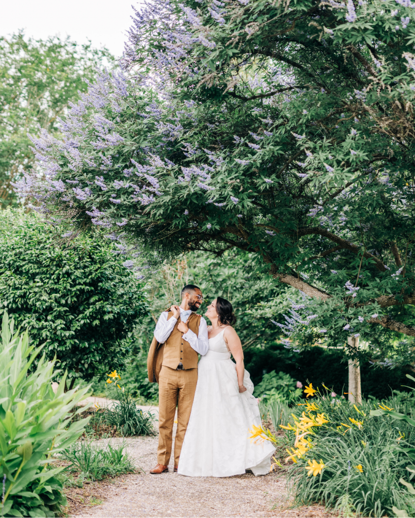Bride and groom stand beneath blooming purple trees along a garden pathway at Daniel Stowe Botanical Garden, creating romantic outdoor wedding portraits featured by Wed Society Charlotte.