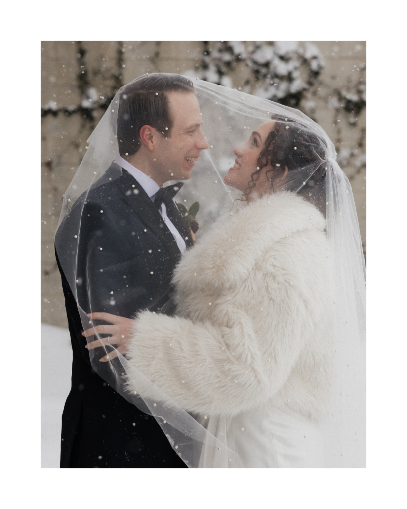 Bride and groom smile beneath a cathedral veil while snow falls during winter wedding portraits in Charlotte, North Carolina.