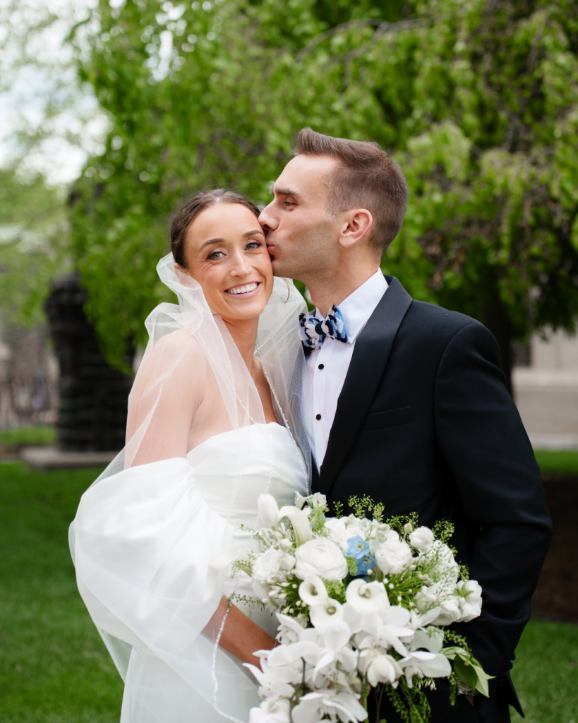 Groom kissing bride's cheek outdoors with white and blue wedding bouquet in foreground
