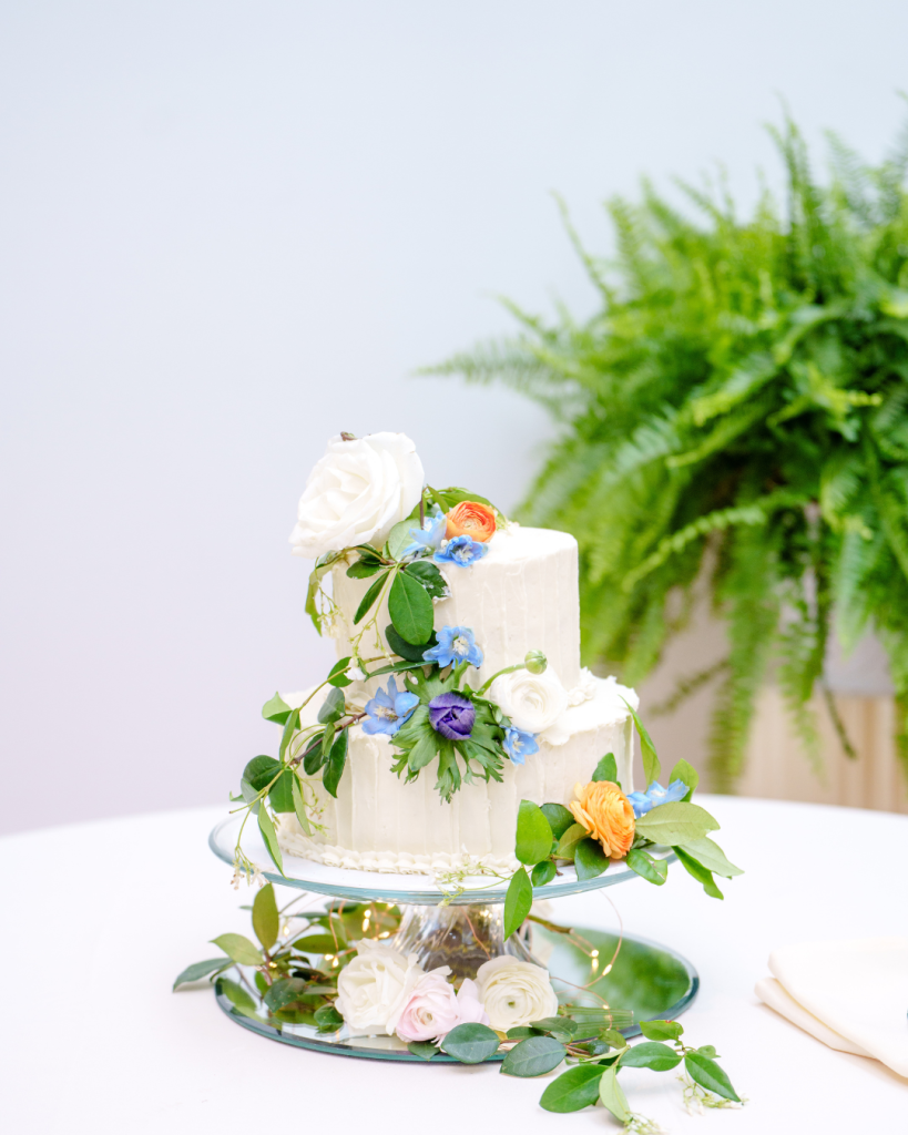 Two-tier white wedding cake decorated with fresh blue, purple, and orange flowers