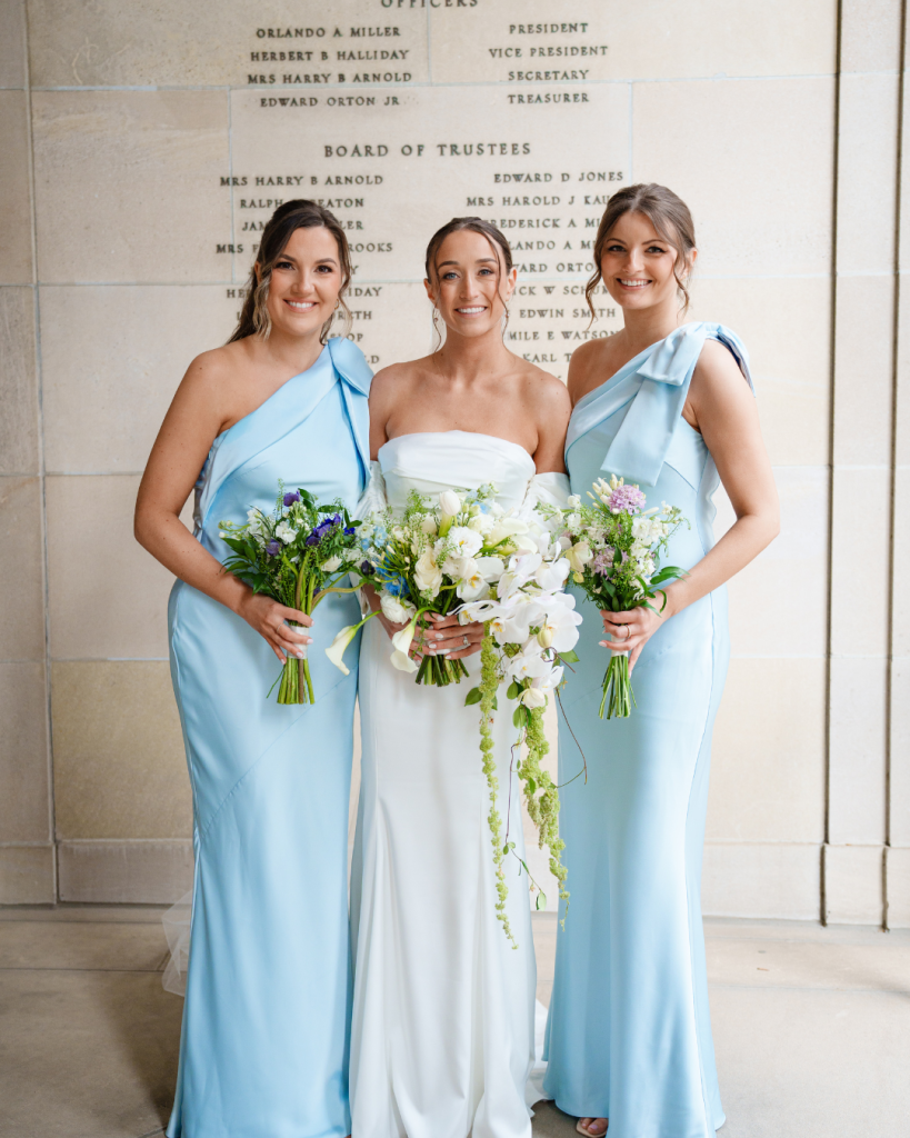 Bride with bridesmaids in light blue one-shoulder gowns holding white and blue floral bouquets