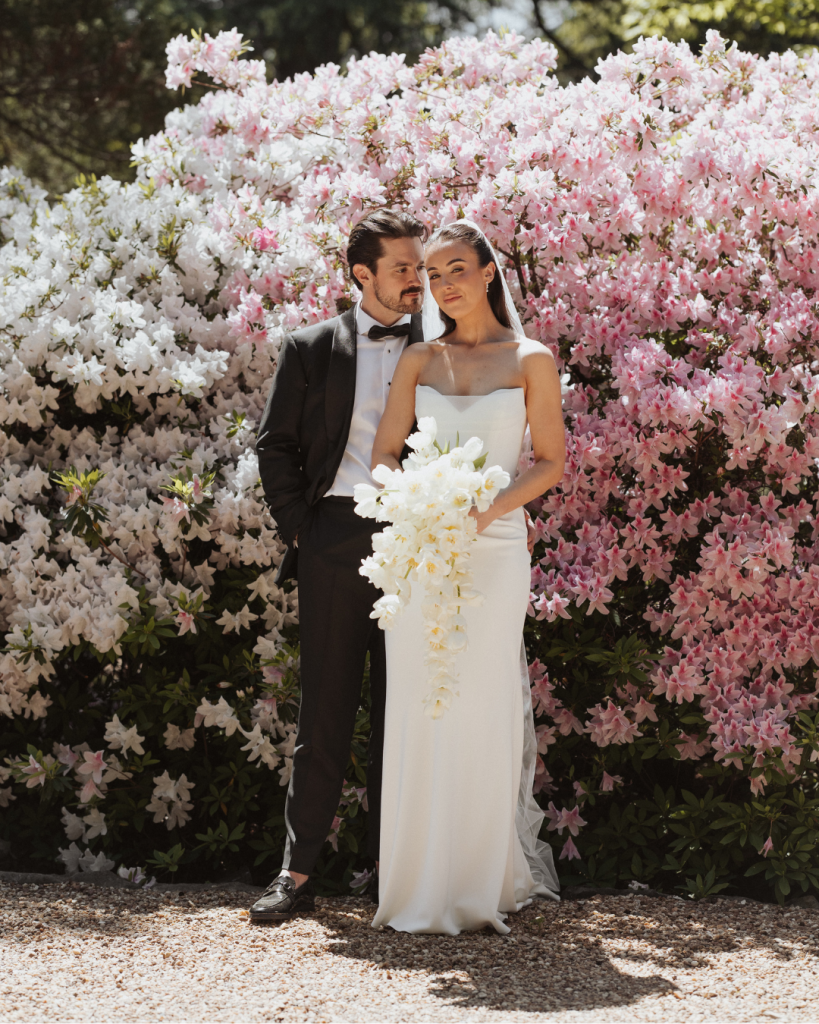 Bride and groom pose beneath blooming pink and white azaleas at The VanLandingham Estate in Charlotte, North Carolina. The bride wears a modern white gown and carries a cascading tulip bouquet during their Wed Society Charlotte featured wedding.