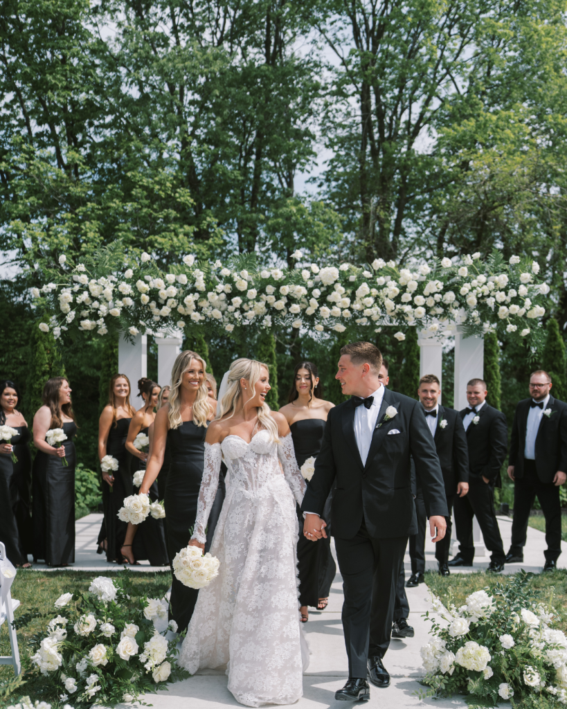 Sierra and Logan pose between black garden gates covered in white flowers at White Willow Meadows near Columbus.