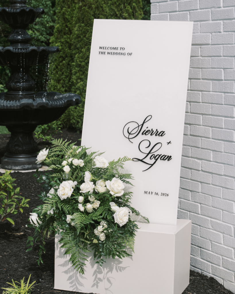 Modern white welcome sign for Sierra and Logan’s May wedding displayed with white roses and greenery.