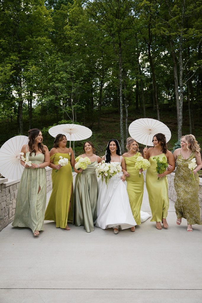 Bride with her bridesmaids dressed in olive and moss green holding white parasols walking across a bridge at Knotting Hills Wedding Venue & Resort.