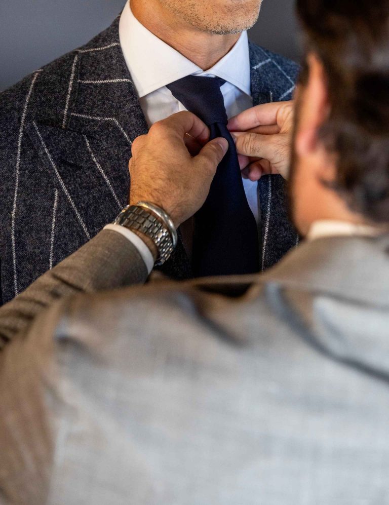 Professional clothier adjusting a navy tie on a client wearing a custom windowpane suit.