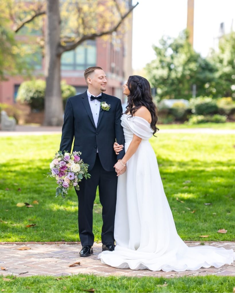 Groom in navy tuxedo kissing bride's cheek as she smiles, holding a bouquet of purple and white flowers with greenery, outdoors on a lawn.