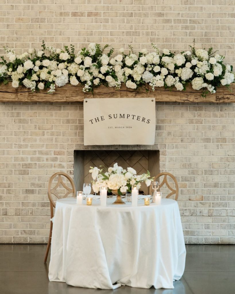 Sweetheart table set against a stone fireplace, topped with a "The Sumpters" sign and framed by a lush white floral garland