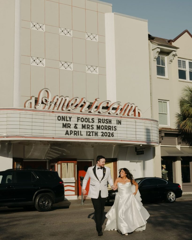Vintage theater marquee wedding venue with a bride in a satin gown and a groom in a white tuxedo jacket.
