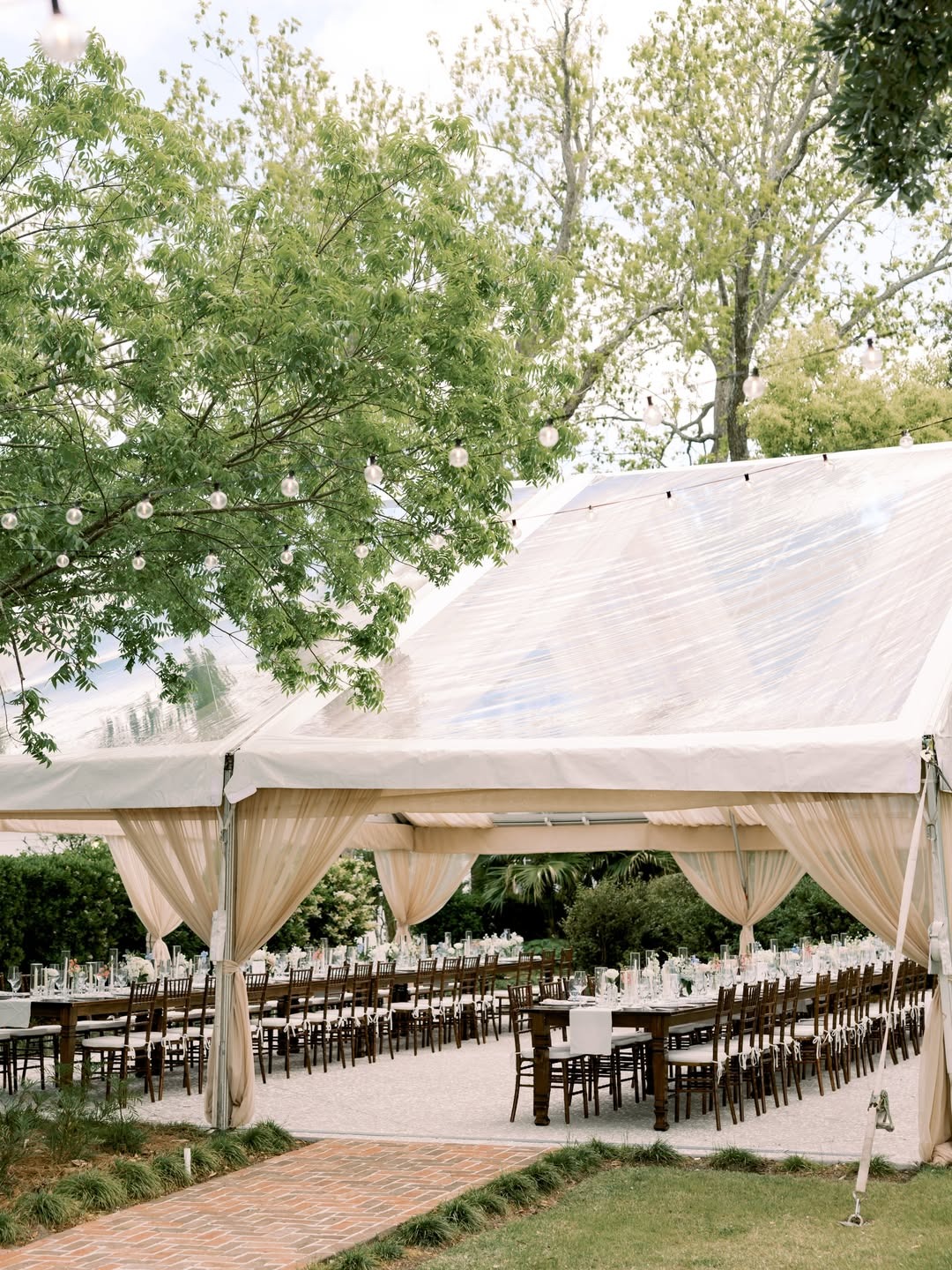 Clear tent wedding reception layout with long wooden tables, neutral drapery, and outdoor lighting ideas.