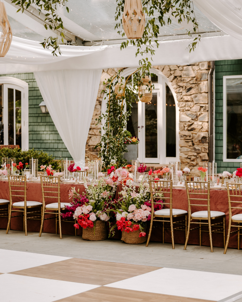 Long reception tables with blush velvet linens, colorful spring floral arrangements, candlelight, and gold chairs beneath a draped clear top tent at Van Landingham Estate in Charlotte, NC featured by Wed Society Charlotte.