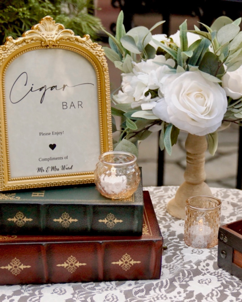 Vintage inspired cigar bar display with gold frame, candles, florals, and decorative books at the wedding reception.