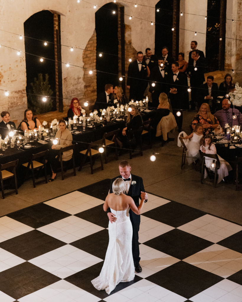 Bride and groom sharing their first dance beneath café lights and chandeliers surrounded by guests at The Providence Cotton Mill wedding reception.