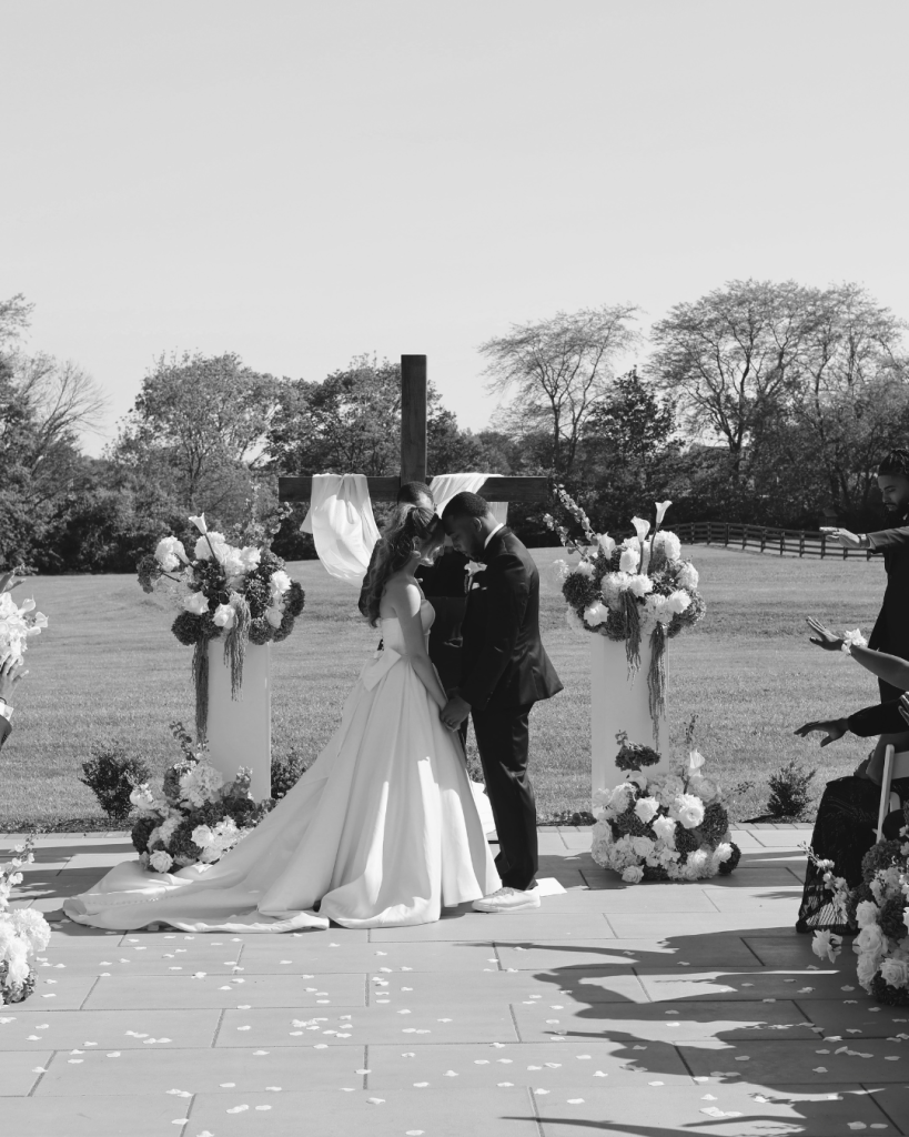 TreVeyon and Kenzie share a kiss beneath a floral-adorned cross as guests celebrate during their outdoor ceremony at Twin Run Farm.