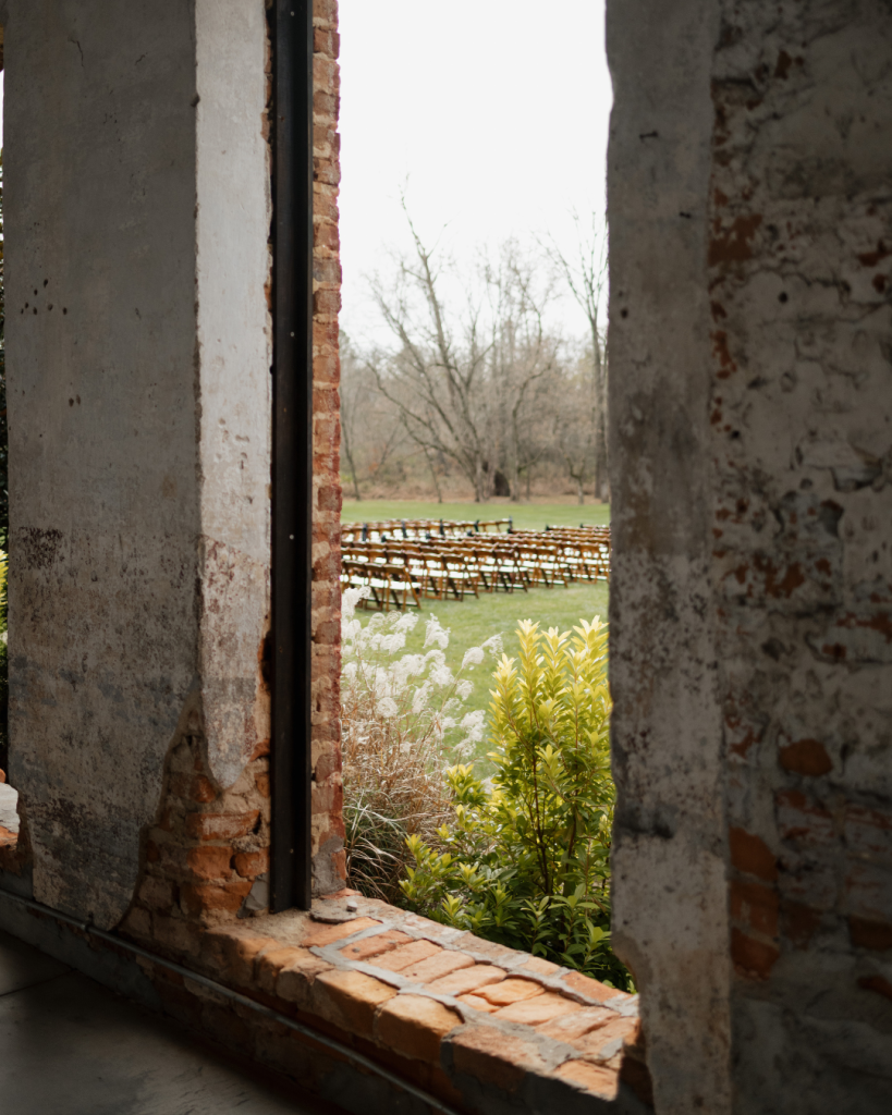 Ceremony view framed through historic brick windows overlooking outdoor seating at The Providence Cotton Mill near Charlotte.