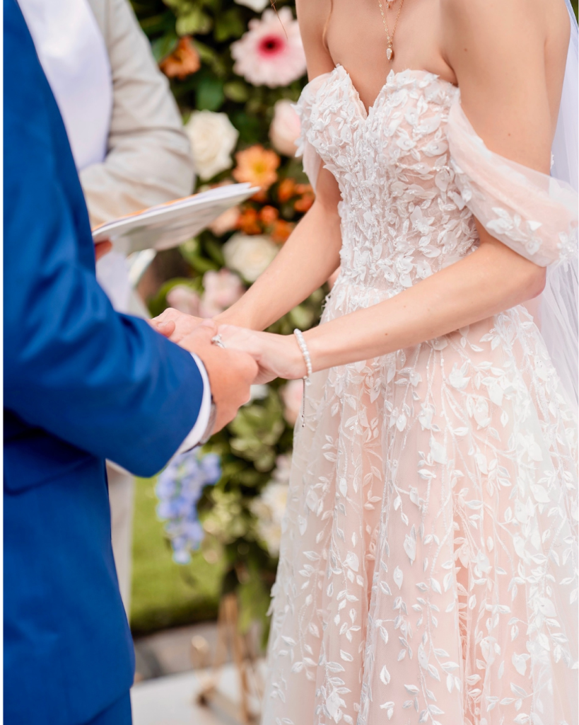 Close up of the bride and groom holding hands during their wedding ceremony beneath a colorful floral installation.