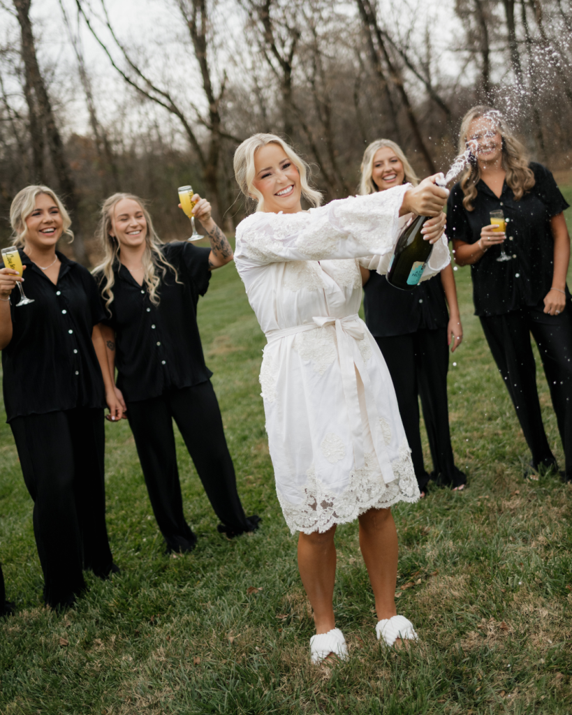 Bride celebrating with bridesmaids while opening a champagne bottle outdoors before her Charlotte area wedding.