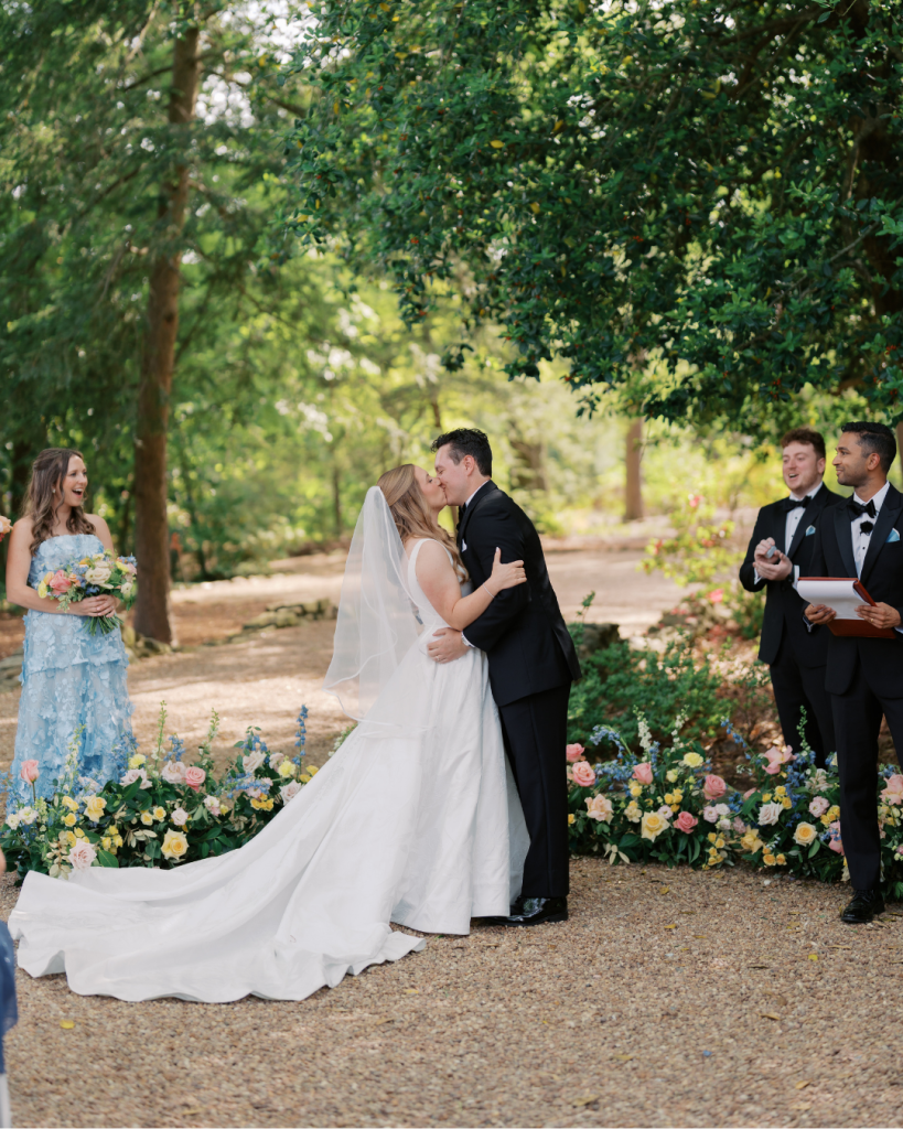 Bride and groom share their first kiss during an outdoor garden ceremony surrounded by colorful floral arrangements at Van Landingham Estate.