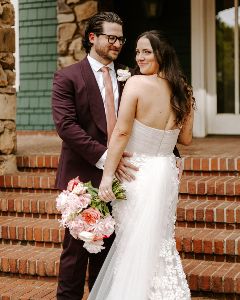 Bride and groom pose together on brick steps outside Van Landingham Estate with colorful bouquet and burgundy suit.