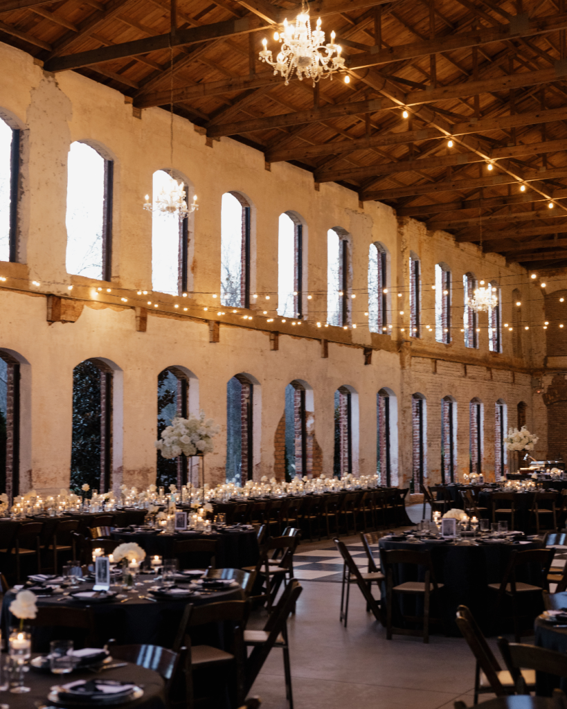 Wide view of candlelit reception with black linens, chandeliers, and historic architecture at The Providence Cotton Mill near Charlotte, NC.