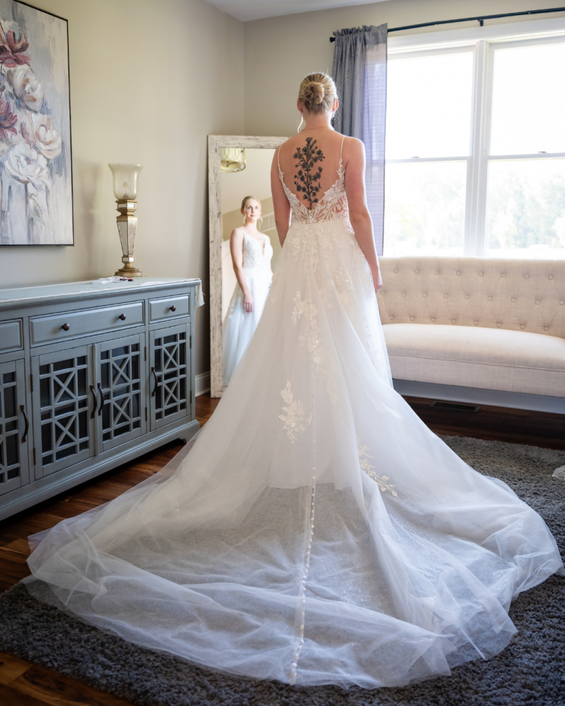 Back view of a bride’s lace wedding gown with a floral illusion back, button detail and long train reflected in a mirror.