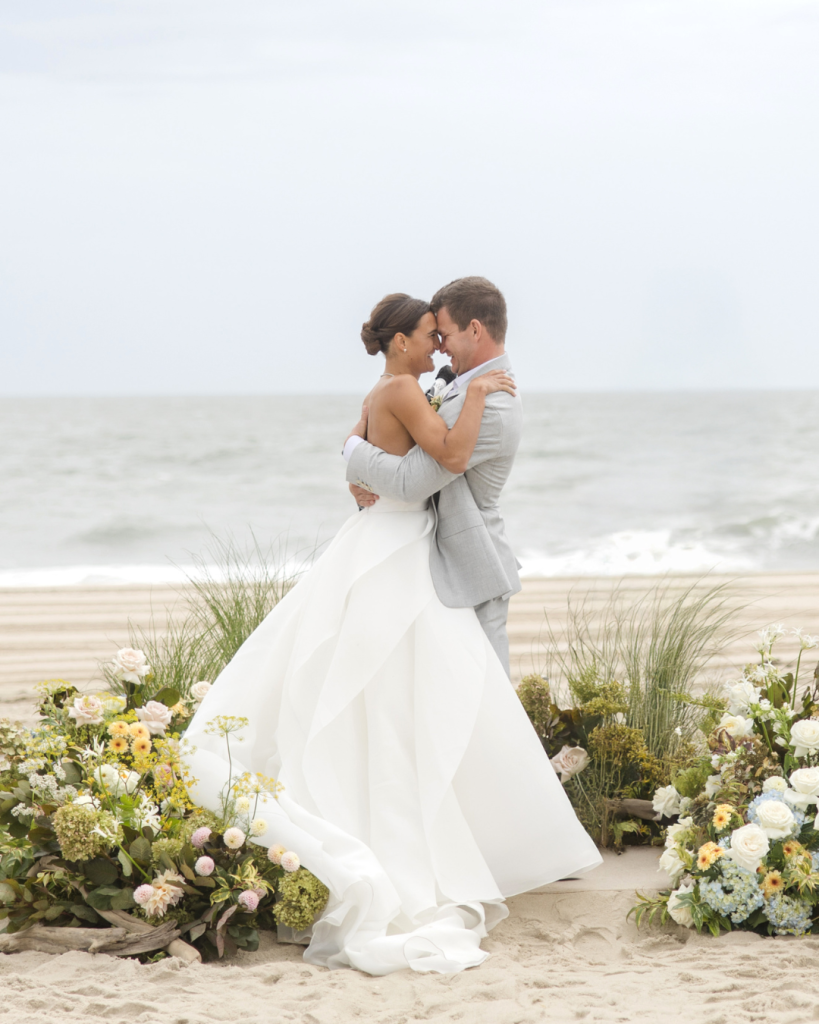 Bride and groom embracing at their beachfront wedding ceremony in Rehoboth Beach, Delaware with ocean waves and coastal floral arrangements behind them.