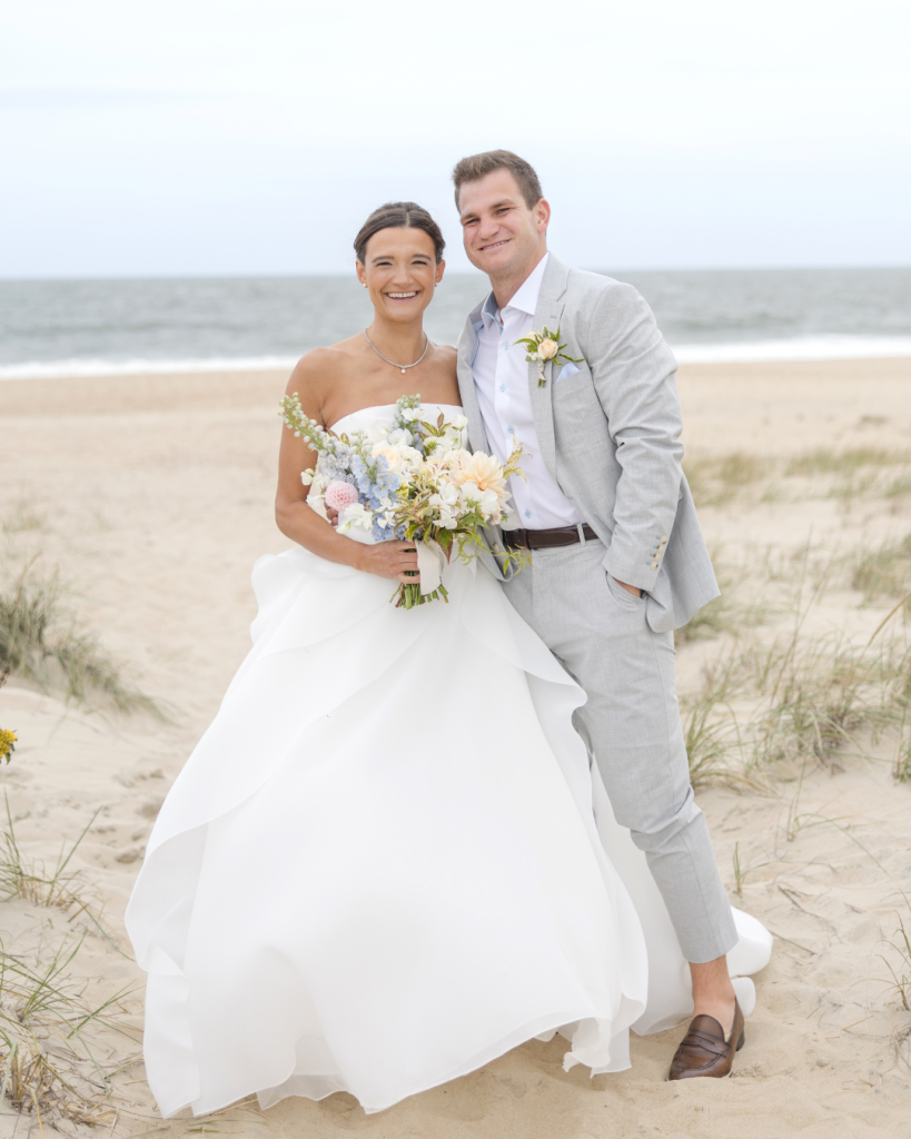 Bride and groom posing on the sandy shoreline during their Rehoboth Beach, Delaware wedding with the Atlantic Ocean and coastal dunes in the background.