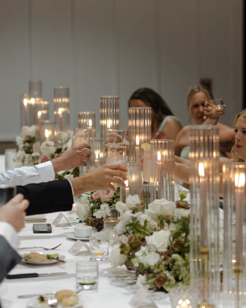 Wedding guests raise their glasses around a candlelit reception table decorated with romantic white and blush florals.