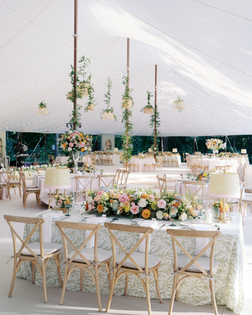 Tented wedding reception with a green-patterned head table, pastel floral arrangements, light wood chairs, hanging greenery and string lights.