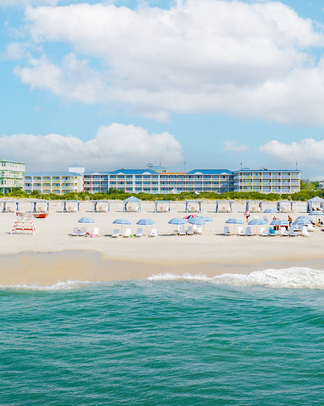Sunny beachfront view of La Mer Beachfront Resort with blue-and-white striped umbrellas, lounge chairs, and turquoise ocean waves.