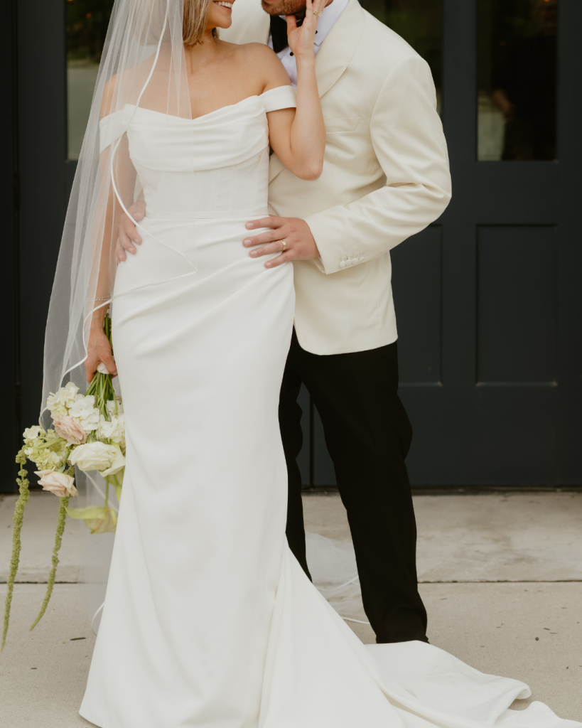 Emily and Joshua pose in their wedding attire outside The Ellis in Columbus, with Emily holding an ivory, blush, and green bouquet.