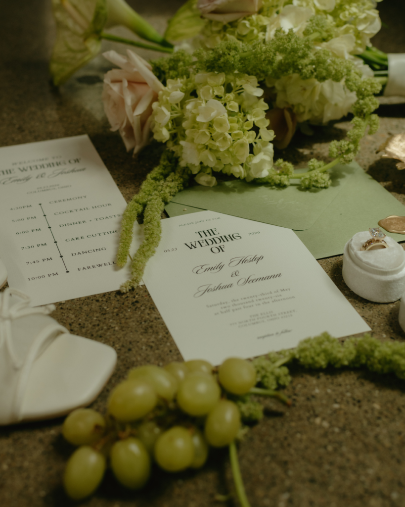 Emily and Joshua’s wedding invitation suite displayed with pale green envelopes, spring flowers, wedding rings, ribbon, and green grapes.